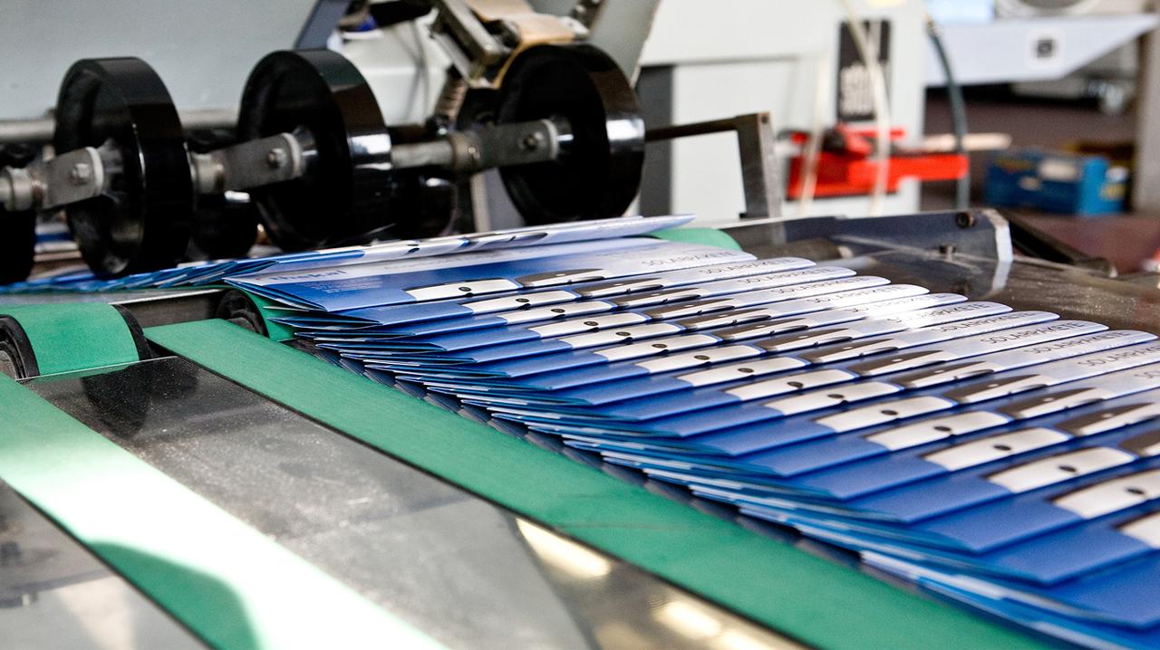Blue printed booklets stacked on a conveyor belt in the Cal Poly Print & Copy production area.